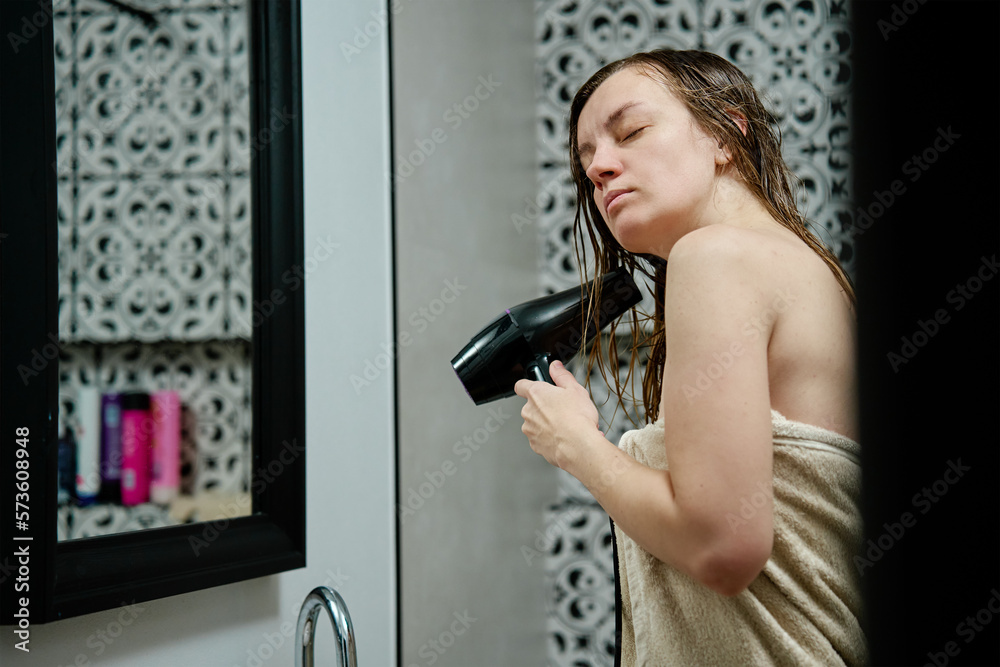 Woman wrapped in towel blow drying hairs standing in bathroom. Female morning hygienic routine and hair care