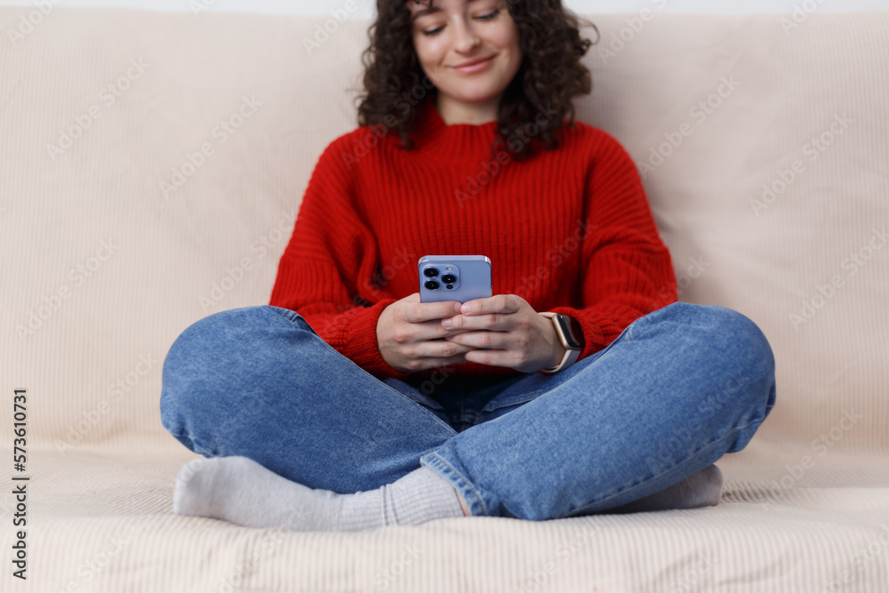 Cheerful young female sitting on couch and typing a message in a smart phone. White girl using modern blue mobile phone