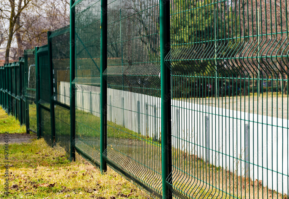 green painted metal wire fence between steel circular posts. transparent screen. welded wire mesh. green grass in the background. sport field beyond. green painted metal wire fence between steel circular posts. transparent screen. welded wire mesh. green grass in the background. sport field beyond.
