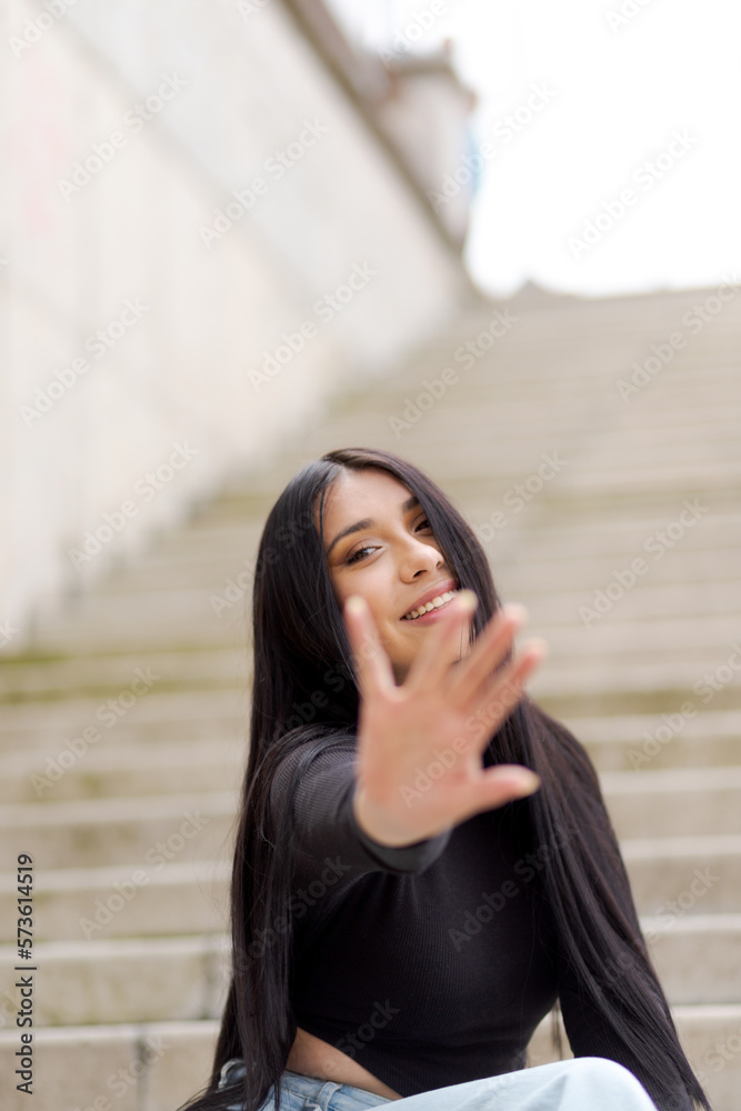 Young woman blocking camera with hand. Happy young man smiling at camera cheerfully while sitting outdoors on street stairs. Vibrant young woman wearing long straight black hair.