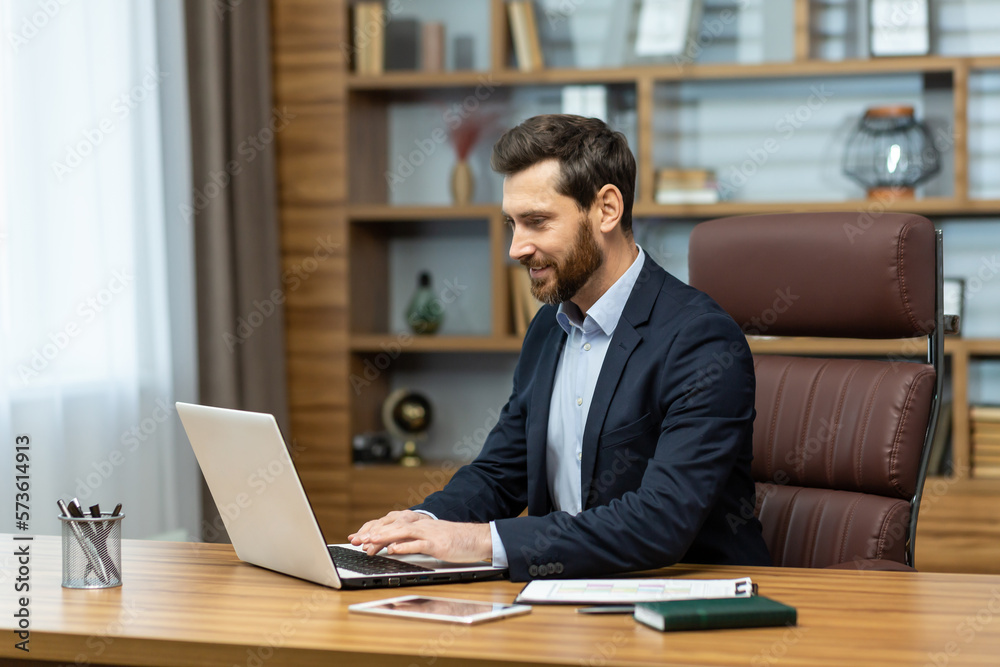 Obraz premium Successful mature businessman lawyer working inside classic modern office, modern man satisfied with work working with laptop at workplace, boss in business suit satisfied with achievement results.