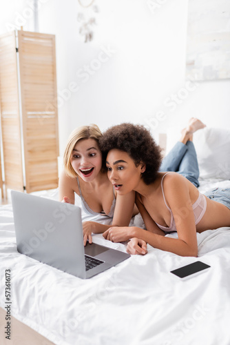 amazed blonde woman pointing at laptop near african american friend and smartphone with blank screen on bed.