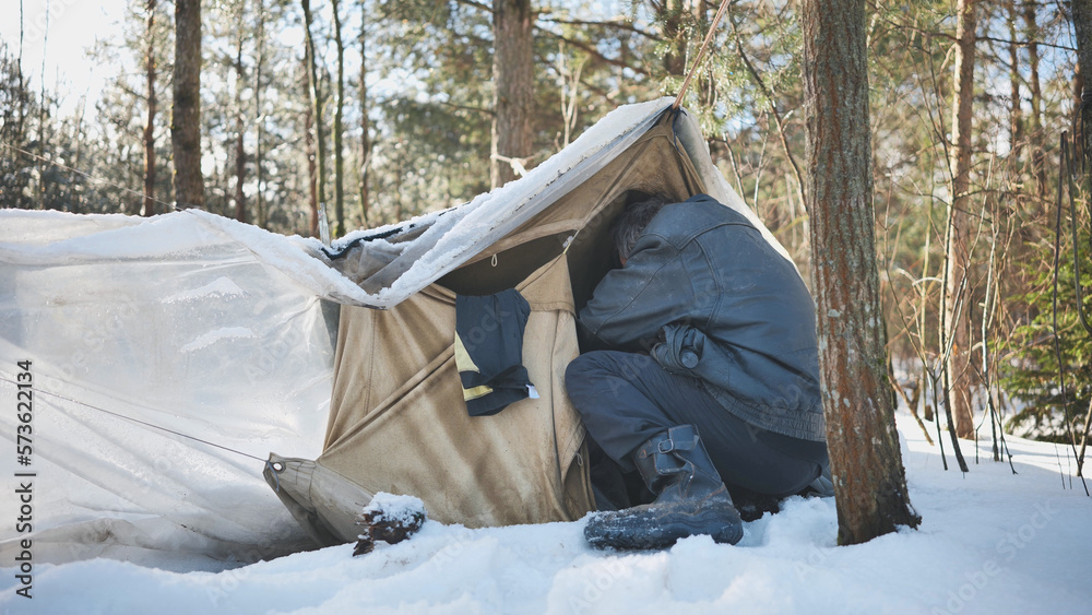A homeless man climbs in and out of a tent in the woods in winter. - obrazy, fototapety, plakaty 