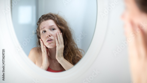 Anxious young woman look in the mirror worried about wrinkle or acne on unhealthy skin, upset unhappy millennial female examine squeeze pimple on face in the bedroom, cosmetology, skincare concept photo