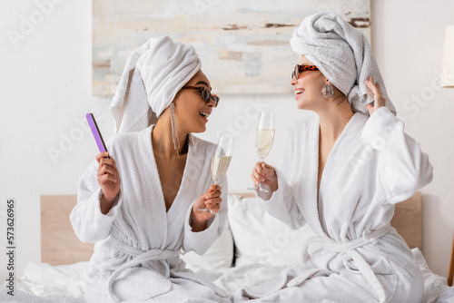 happy interracial women in terry robes and stylish sunglasses holding champagne and looking at each other in bedroom.