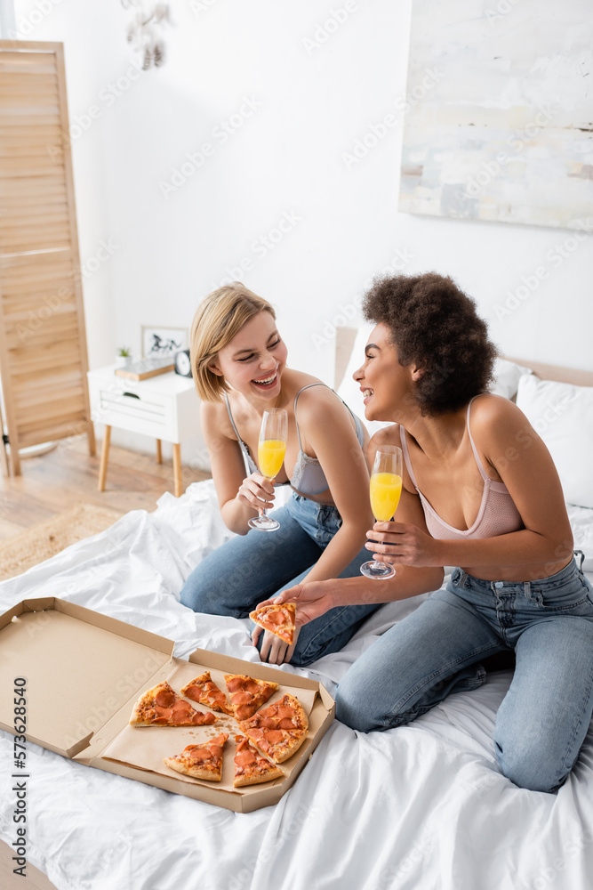 cheerful interracial women in bras and jeans sitting with cocktails in champagne glasses on bed at home.