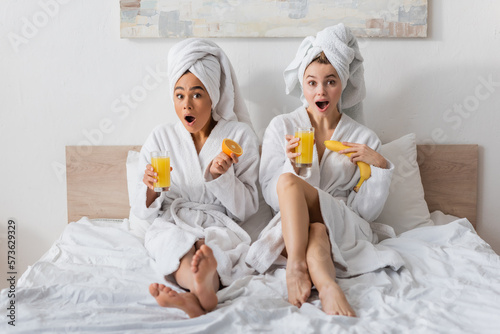 full length of amazed interracial women in white robes and towels holding fruits and orange juice while looking at camera on bed.