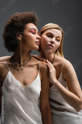 sensual interracial women with colorful makeup posing in strap dresses and silver necklaces isolated on grey.