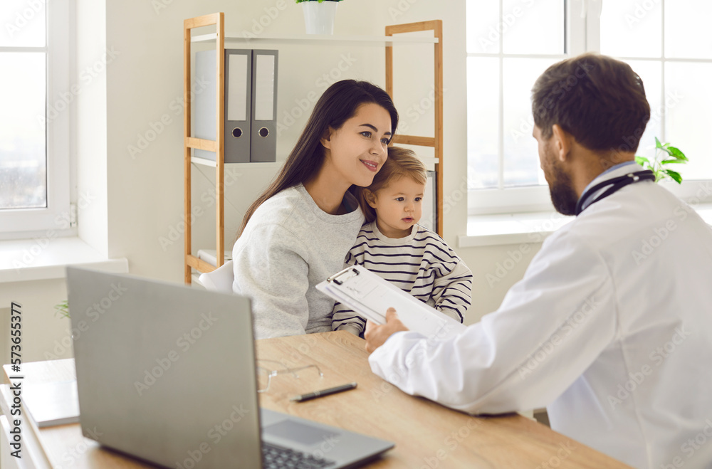 Male pediatrician examining baby and filling form at consultation. Young mother with toddler baby talking to doctor GP and listening recommendations at meeting in medical office Male pediatrician examining baby and filling form at consultation. Young mother with toddler baby talking to doctor GP and listening recommendations at meeting in medical office