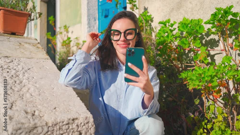 Woman in glasses using smartphone outdoor sitting stairs looking on device screen and smile. Happy female millennial wears casual clothes sits near building reads message on smartphone enjoys summer