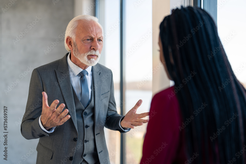 Furious senior businessman with gray hair and beard in conflict with young black haired businesswoman. Senior businessman showing anger, potential violence at work, bullying young coworker.