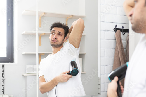 Man sweating excessively and trying to dry his armpits with hair dryer in front of mirror in the bathroom