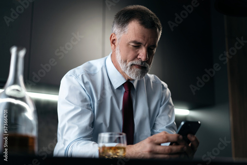 Mature businessman with glass of whiskey using smartphone while sitting in the kitchen.
