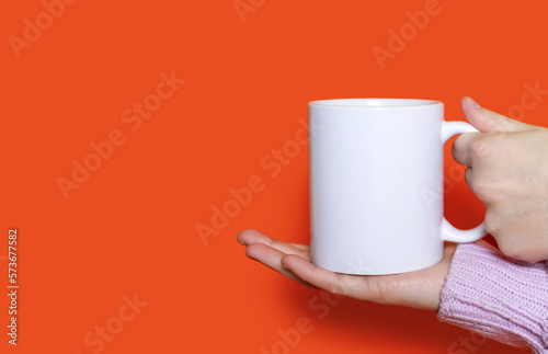 A woman's hand holds a white cup with a hot drink on an orange background. The hand of a woman dressed in a pink sweater holds a ceramic cup on an orange background. Free space for text