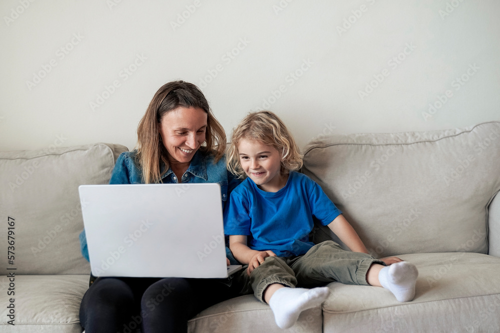 Happy mother and kid son watching videos in computer laptop inside living room at home - Focus on mom face - Family love concept Happy mother and kid son watching videos in computer laptop inside living room at home - Focus on mom face - Family love concept