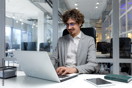 Cheerful and successful businessman inside office working with laptop, bearded man smiling and typing on laptop keyboard, programmer coding software in modern room.