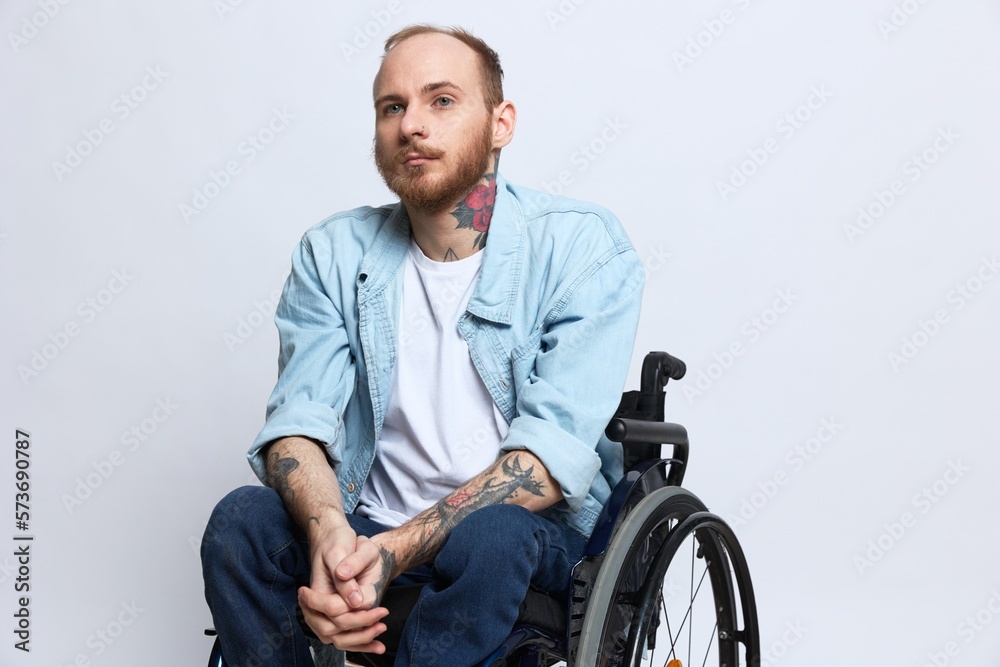 A man in a wheelchair looks thoughtfully at the camera, copy space, with tattoos on his arms sits on a gray studio background, the concept of health a person with disabilities, a real person A man in a wheelchair looks thoughtfully at the camera, copy space, with tattoos on his arms sits on a gray studio background, the concept of health a person with disabilities, a real person