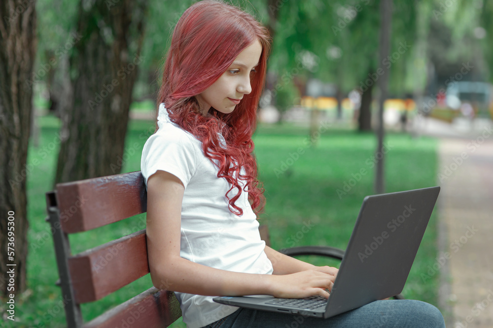 Young girl teenager or student in casual clothing with red hair sitting on a bench in spring or summer park with laptop, looking at her computer and typing, studying, communicating or working online Young girl teenager or student in casual clothing with red hair sitting on a bench in spring or summer park with laptop, looking at her computer and typing, studying, communicating or working online