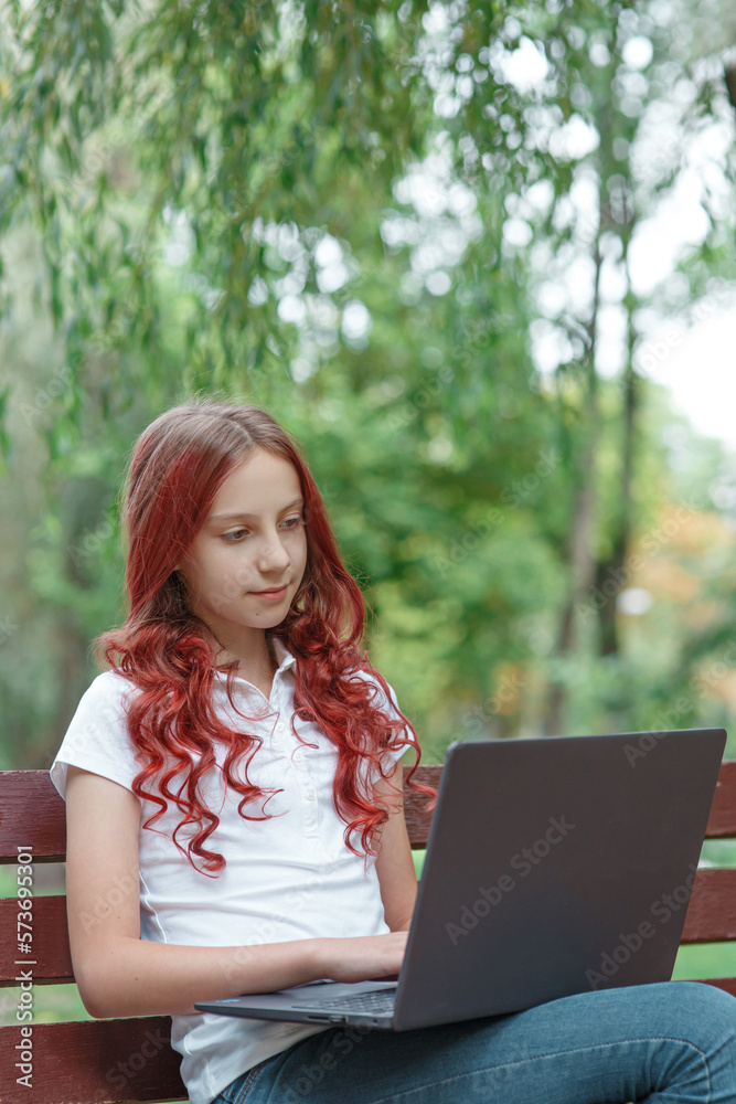 Young dreaming girl teenager or student in casual clothing with red hair sitting in spring or summer park with laptop with trees behind, looking at her computer and typing, studying or working online Young dreaming girl teenager or student in casual clothing with red hair sitting in spring or summer park with laptop with trees behind, looking at her computer and typing, studying or working online