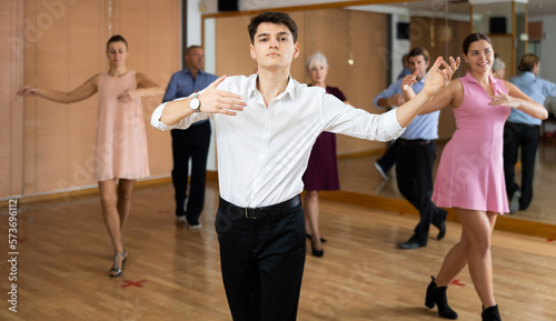 Dynamic young man practicing ballroom dance pose in training hall with other persons during dancing-classes