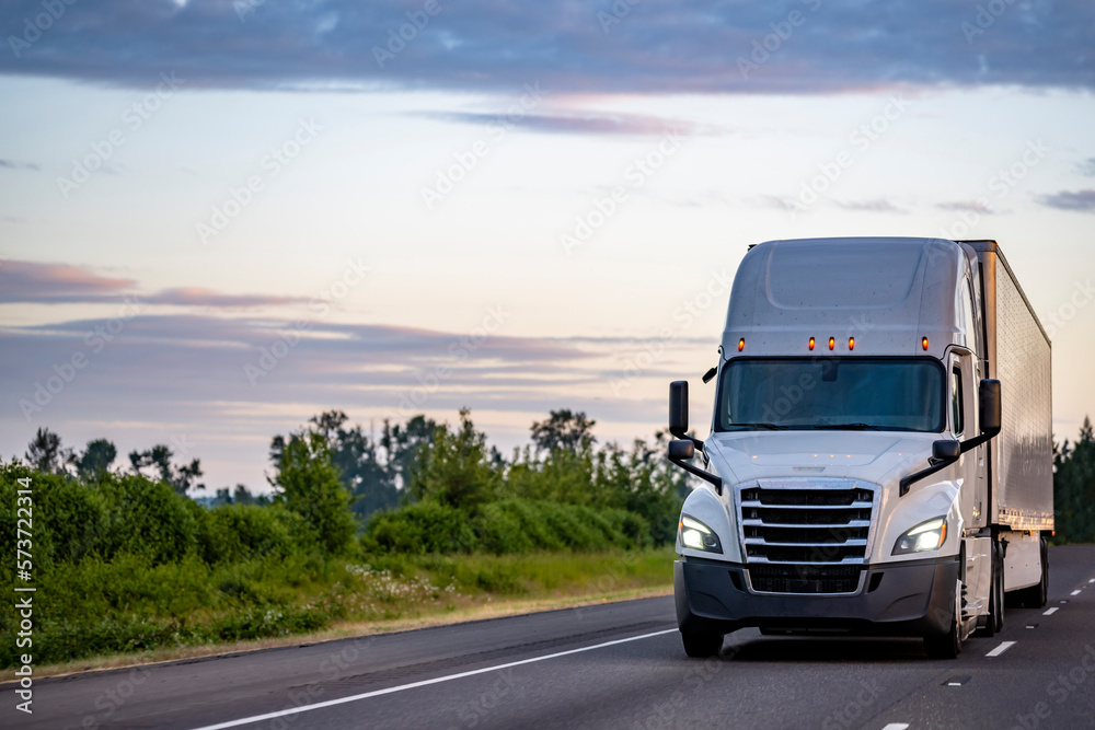 Bonnet big rig industrial white semi truck with turned on headlights transporting cargo in dry van semi trailer driving on the highway road at twilight time