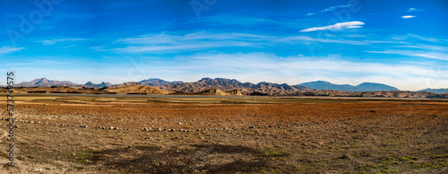 A panorama view ofLake Grassmere farmland with a backdrop of the lake and the rugged mountain range under a blue cloudy sky