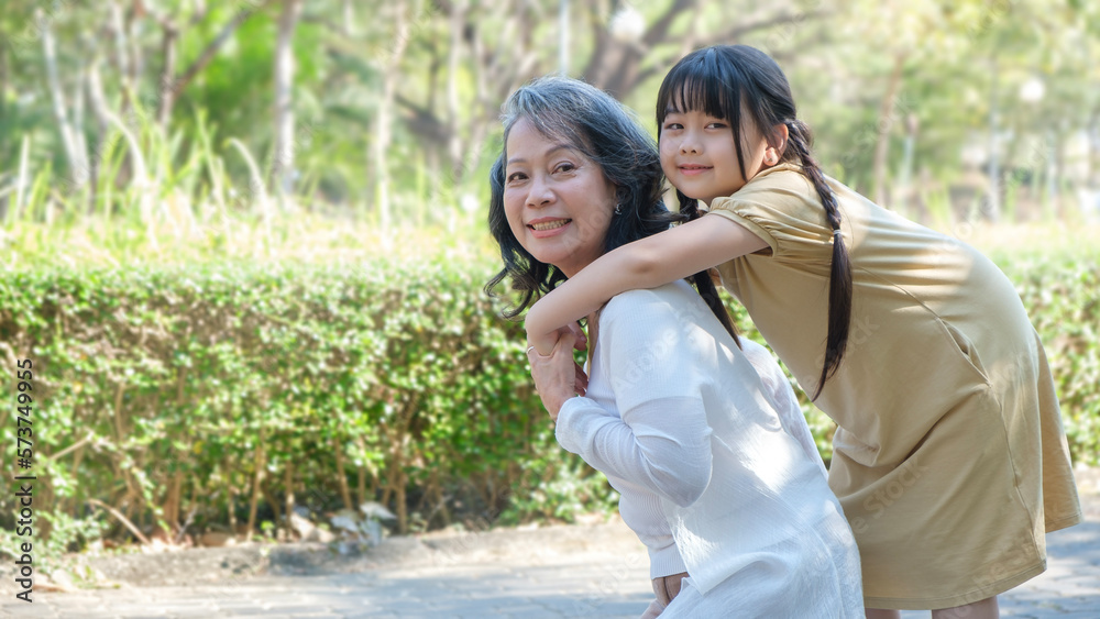 Naklejka premium Smiling middle age grandmother giving piggyback ride to cute little granddaughter during walking in the park.