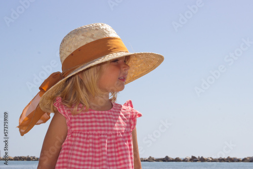 Φωτογραφία Little blond girl in straw hat and sundress stay at the beach with sea on background,portrait of european toddler,adorable kid looking forward