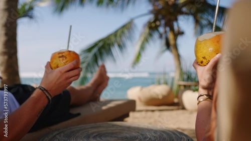 Closeup shot of fresh king coconuts with exotic background. Tourists holding two cocnuts with straws, clink them and drink. Young couple enjoys cool cocnut water at tropical vacation by sea close up.