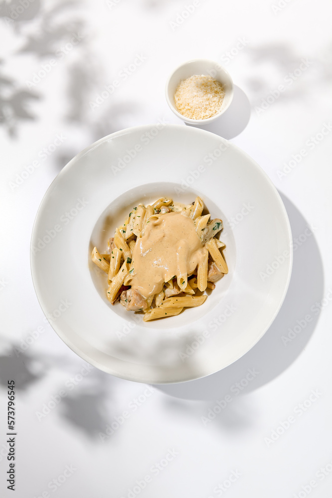 Italian pasta penne with chicken and mushrooms in creamy sauce. Mushroom and chicken pasta on white plate with shadows of sunlight. Summer italian lunch Penne with mushroom sauce on light background