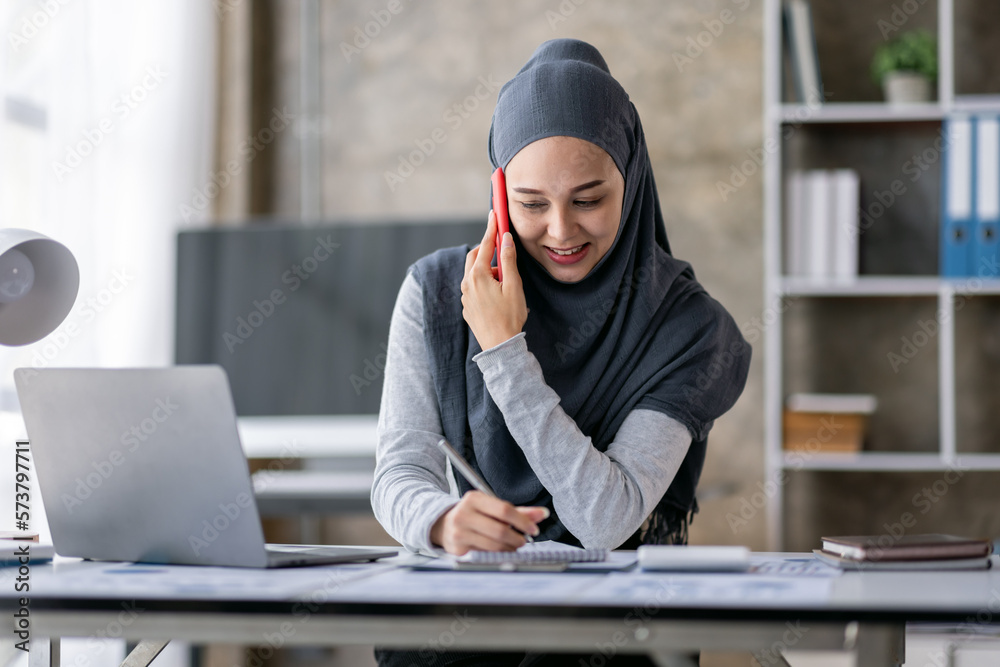 Cheerful business Asian muslim woman freelancer making telephone call share good news about project working in office workplace, business finance concept.	.