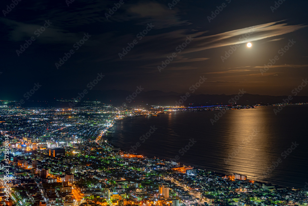 Fototapeta premium Hakodate City night view from Mt. Hakodate observatory, big bright moon light up the sea, golden reflection on surface. Famous scenic spot in the world. Hokkaido, Japan