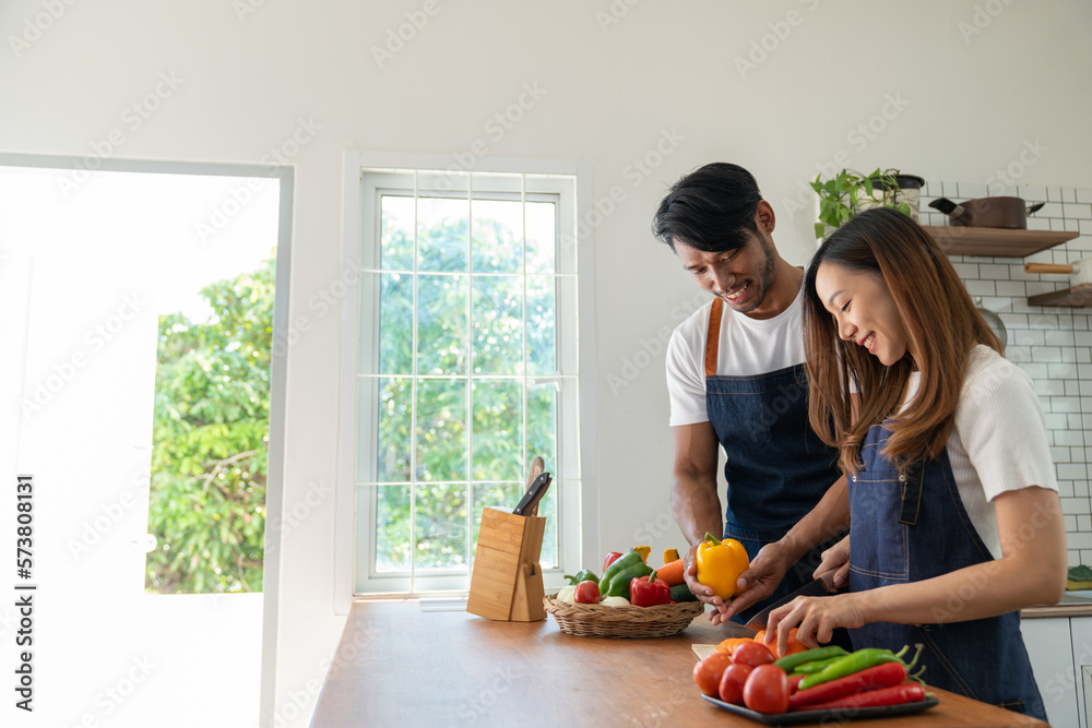 Young Asian couple wearing apron cooking healthy salad in the kitchen together picking out ingredients for the online cooking class. lifestyle and cooking concept.