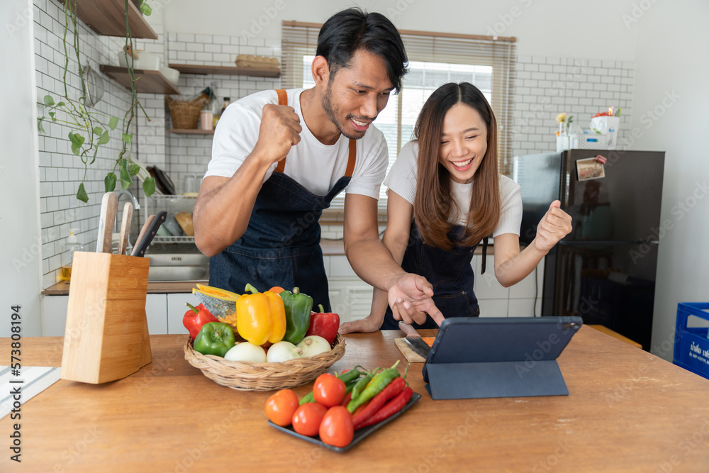 Young Asian couple wearing apron learning how to choose ingredients such as fruits and vegetables to prepare their morning meal via tablet, online. Lifestyle and healthy cooking concept.