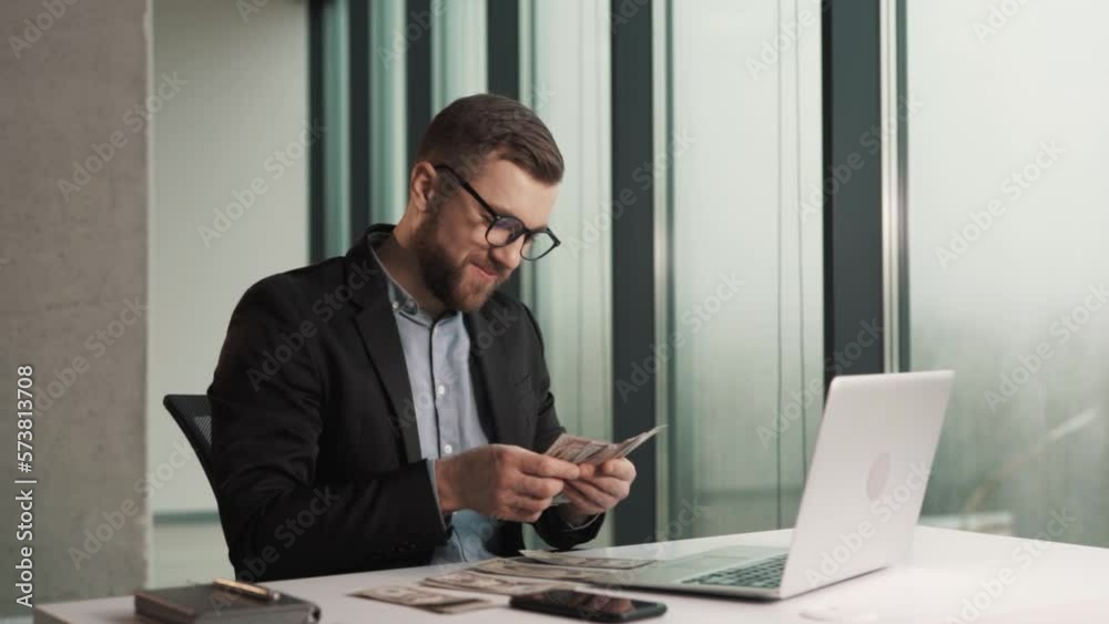 A happy young man in business clothes counts money bills while sitting at a laptop