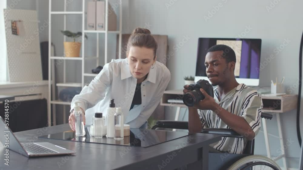 Young African American male photographer in wheelchair photographing cosmetic products in bottles on digital camera, working with female assistant in bright modern photo studio Young African American male photographer in wheelchair photographing cosmetic products in bottles on digital camera, working with female assistant in bright modern photo studio