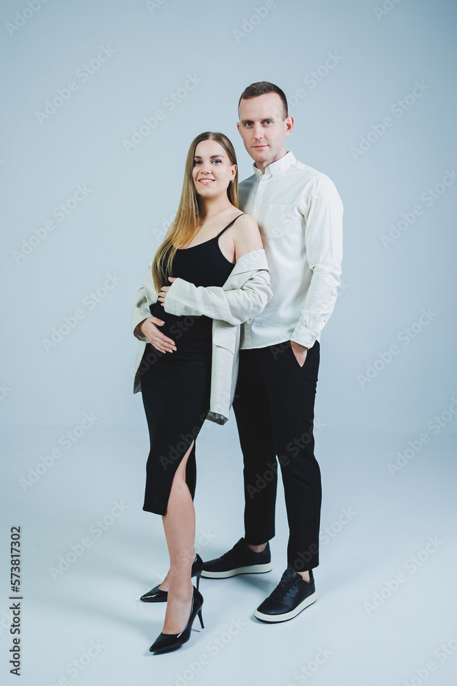 Happy young man standing with his pregnant wife and hugging her belly, isolated on white background. Stylish young people. A man in a shirt and a woman in a black skirt and jacket Happy young man standing with his pregnant wife and hugging her belly, isolated on white background. Stylish young people. A man in a shirt and a woman in a black skirt and jacket