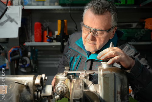 A man in safety glasses and work uniform sets up a grinding machine in his workshop. The grinding machine details in focus. In the background are various working tools on metal shelving for workshop
