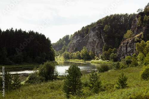 Gentle morning landscape on quiet river with high rocks and lush green pine forest, green meadow on shore in bright golden sunbeams in summer. Sunny scenery for trekking and adventure on outdoors. © finepoints