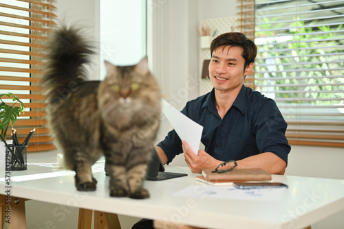 Smiling millennial man with lovely fluffy cat distracted from work in comfortable home office