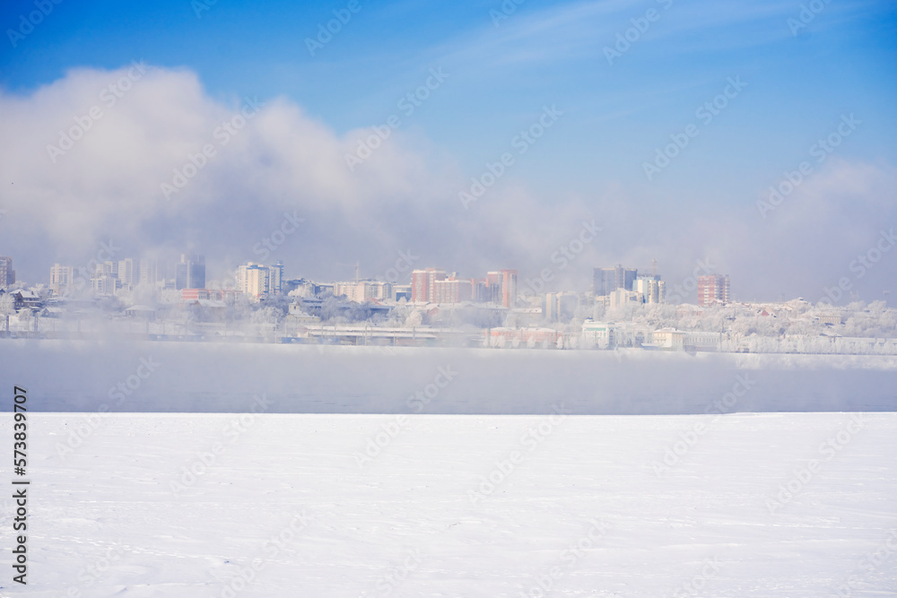 Urban winter landscape with snow on a frozen river against the backdrop of city houses in the fog.