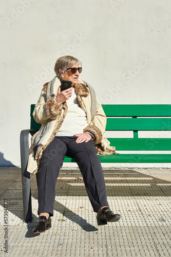 Fototapeta Naklejka Na Ścianę i Meble -  an eighty-year-old woman sunbathes in sunglasses sitting on a green wooden bench in front of a concrete wall while talking on the cellular phone