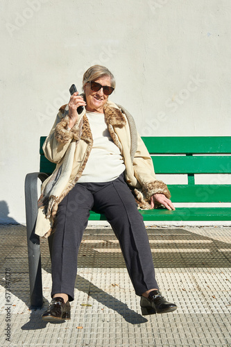 an eighty-year-old woman sunbathes in sunglasses sitting on a green wooden bench in front of a concrete wall while talking on the cellular phone