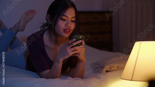 Happy young woman using mobile phone while lying on a bed at night.