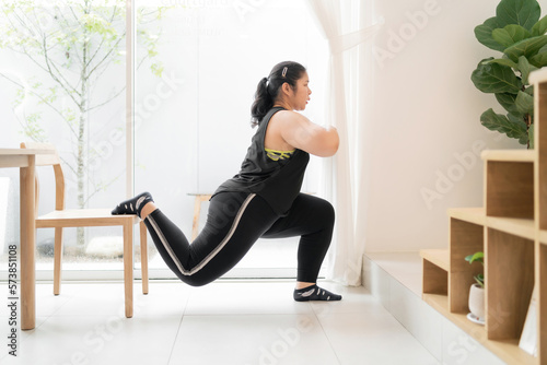 overweight female person workout using chairs in living room single leg lunge. motivated chubby woman trying single leg lunge pose on chairs. fat determined woman back lunge chairs exercising legs