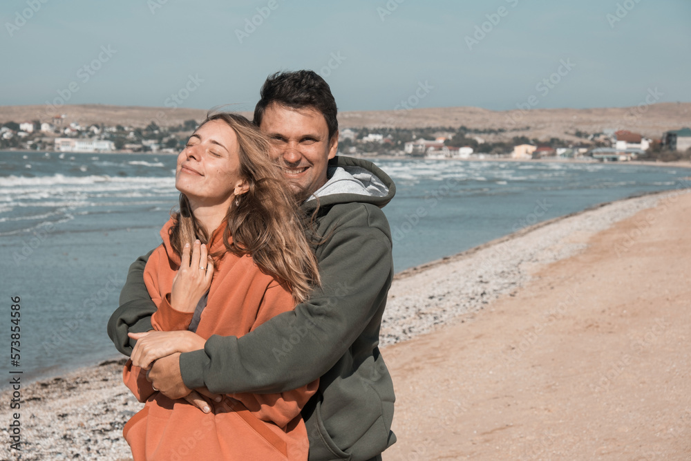The guy and the girl hug and look at each other with love on the beach, against the backdrop of the Blue Sea. Tender relationship of husband and wife. The date of the young couple on Valentine's Day