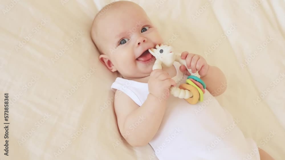 the baby girl is lying on the bed at home playing with a rattle , licking, looking at the camera smiling, top view the baby girl is lying on the bed at home playing with a rattle , licking, looking at the camera smiling, top view