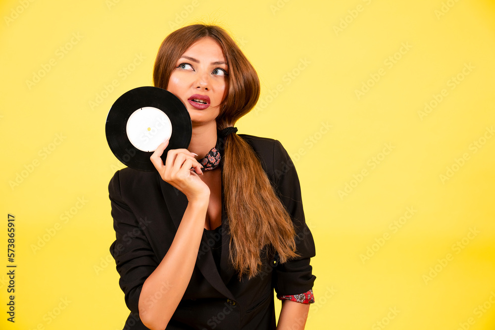Obraz premium girl in a black suit on a yellow background with a record in her hands, music, the girl shows emotions
