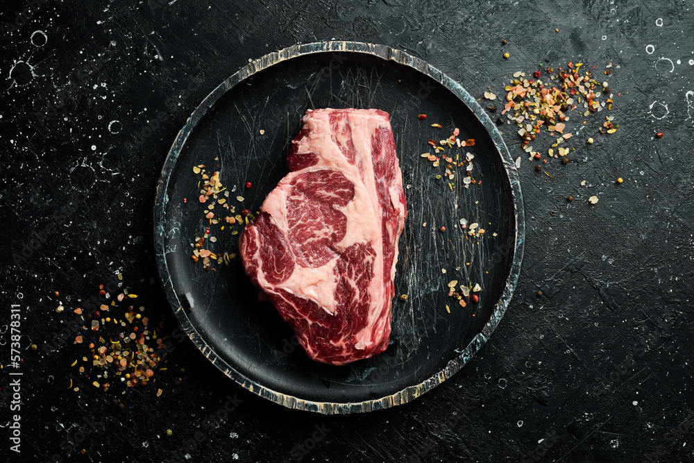 Raw Steak Ribeye Black Angus on a wooden plate ready to be cooked. On a black stone background. Rustic style.