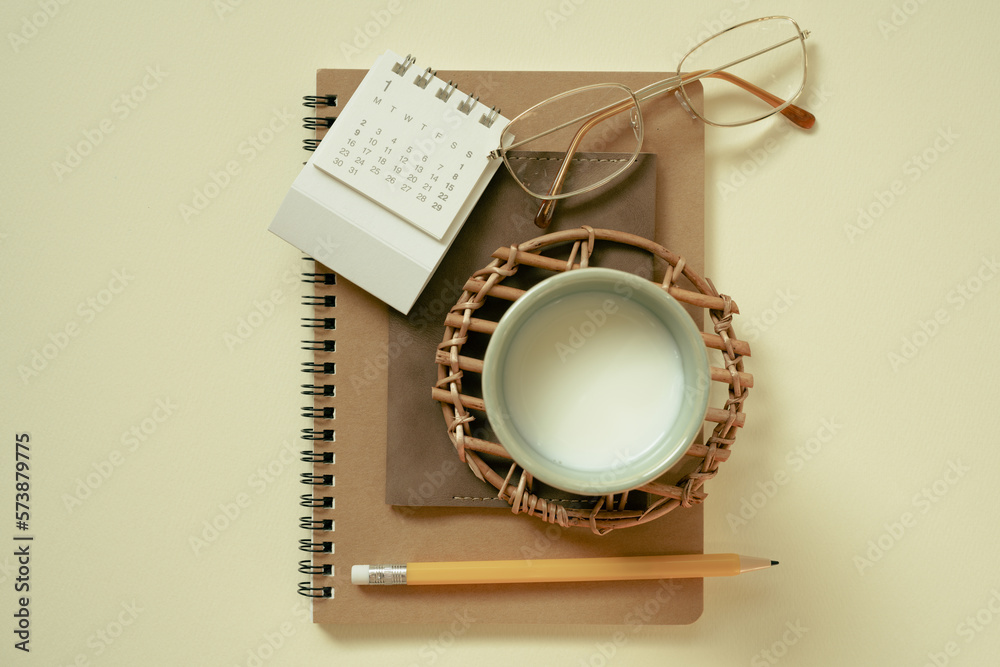 Brown notebook, calendar, eyeglasses, cup of milk, pencil on yellow desk background. flat lay, top view